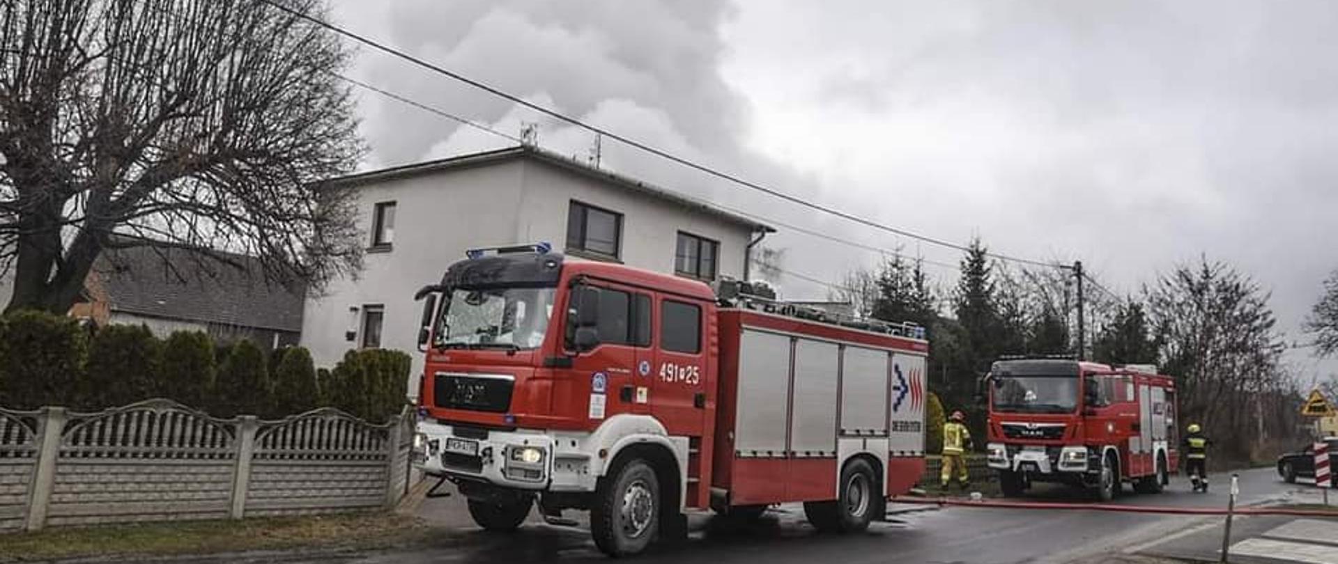 Na drodze przed posesją, na której powstał pożar garaży, znajdują się samochody pożarnicze. Przez jezdnie przebiega linia gaśnicza. Obok samochodów pożarniczych stoi dwóch strażaków w ubraniach specjalnych. Nad budynkiem mieszkalnym widoczne kłęby białego dymu. 