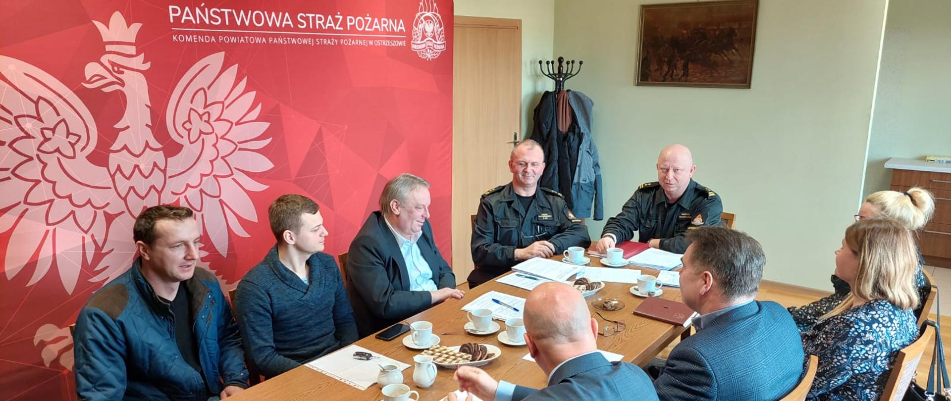 Na zdjęciu osoby siedzące przy stole podczas spotkania. Zdjęcie wykonane w pomieszczeniu. W tle baner Państwowej Straży Pożarnej.
