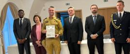 Strażak Centralnej Szkoły PSP trzymający w dłoni otrzymany dokument pozuje do pamiątkowego zdjęcia w towarzystwie kobiety oraz czterech mężczyzn