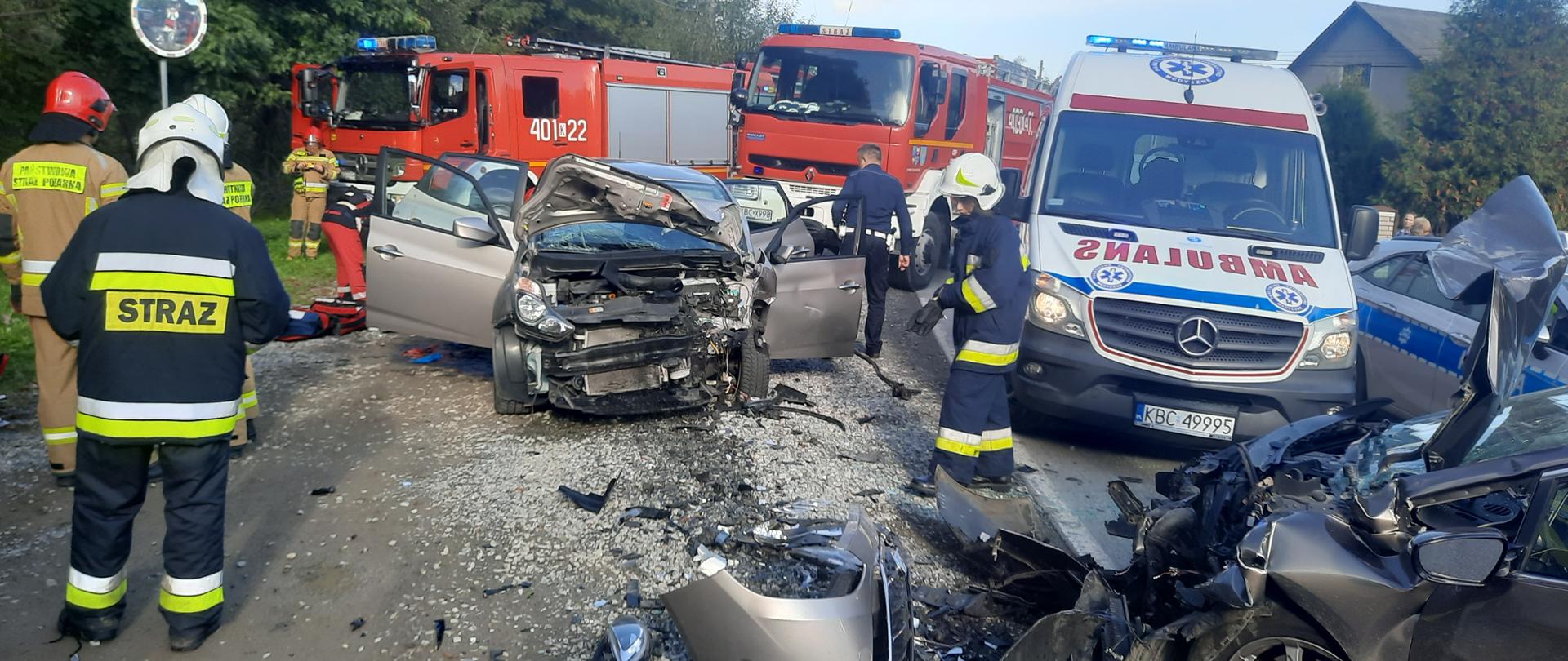 Zdjęcie przedstawia zderzenie dwóch samochodów osobowych. Na zdjęciu widać uszkodzony przód pojazdu osobowego., samochód ma otwarte wszystkie drzwi obok niego widać stojących strażaków ubranych w ubrania specjalne i hełmy. Obok drzwi samochodu widać pochylającego się nad osobą poszkodowaną ratownika medycznego. Po prawej stronie widać część uszkodzonego przodu drugiego pojazdu osobowego. Zanim widać Bok pojazdu Policji oraz przód karetki ZRM. Za pojazdami widać dwa ciężarowe samochody pożarnicze. Za pojazdami widać część budynku mieszkalnego.