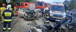 Zdjęcie przedstawia zderzenie dwóch samochodów osobowych. Na zdjęciu widać uszkodzony przód pojazdu osobowego., samochód ma otwarte wszystkie drzwi obok niego widać stojących strażaków ubranych w ubrania specjalne i hełmy. Obok drzwi samochodu widać pochylającego się nad osobą poszkodowaną ratownika medycznego. Po prawej stronie widać część uszkodzonego przodu drugiego pojazdu osobowego. Zanim widać Bok pojazdu Policji oraz przód karetki ZRM. Za pojazdami widać dwa ciężarowe samochody pożarnicze. Za pojazdami widać część budynku mieszkalnego.