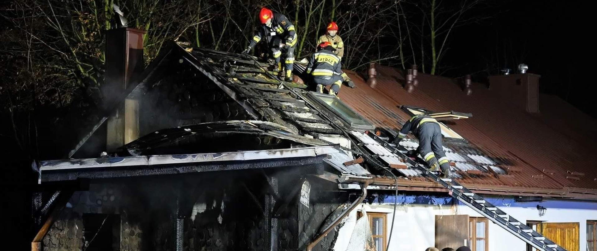Na zdjęciu widoczny jest częściowo spalony budynek jednorodzinny. Spalone są:elewacja ściany szczytowej oraz część dachu. Na dachu strażacy prowadzą prace rozbiórkowe.