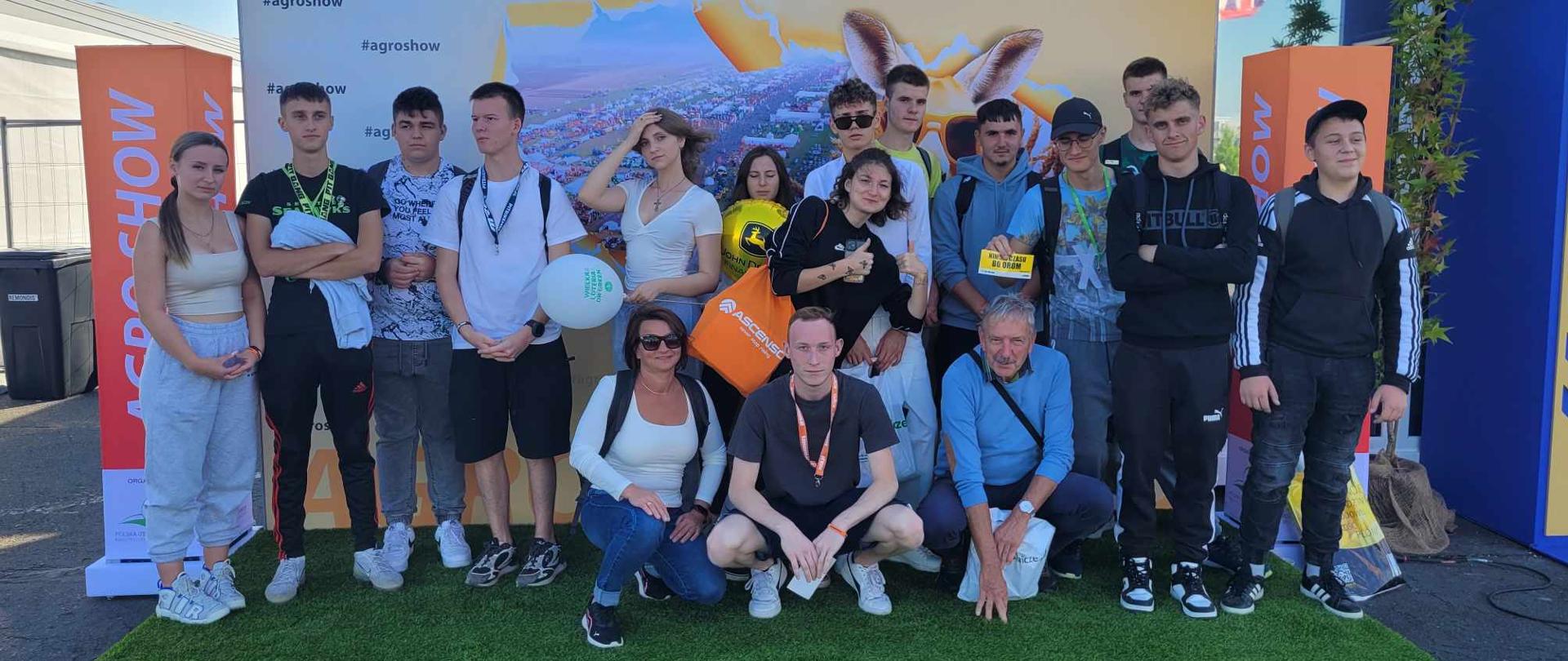 Grupa osób pozująca do wspólnego zdjęcia na tle dużej tablicy informacyjnej z napisem „Agro Show edycja 25”.