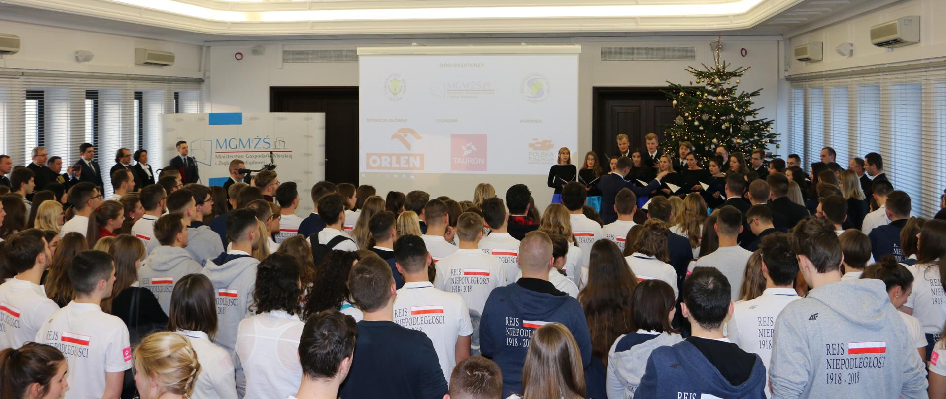 Widok na całą salę po lewej kierownictwo resortu po prawej chór. Na pierwszym planie uczestnicy rejsu widziani od tyłu. 