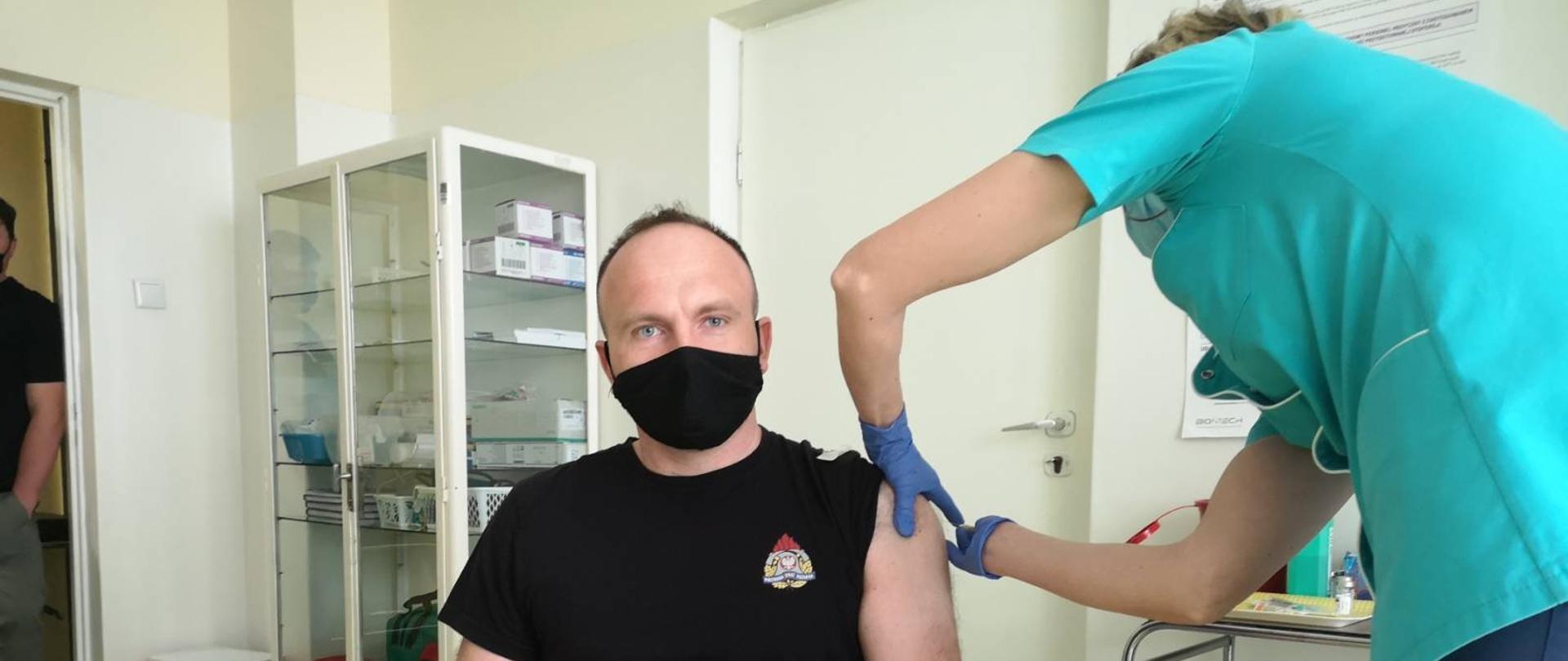 Pielęgniarka w zielonym uniformie szczepiąca strażaka w gabinecie lekarskim. Strażak siedzi na krzesełku w częściowym umundurowaniu koszarowym koloru czarnego.