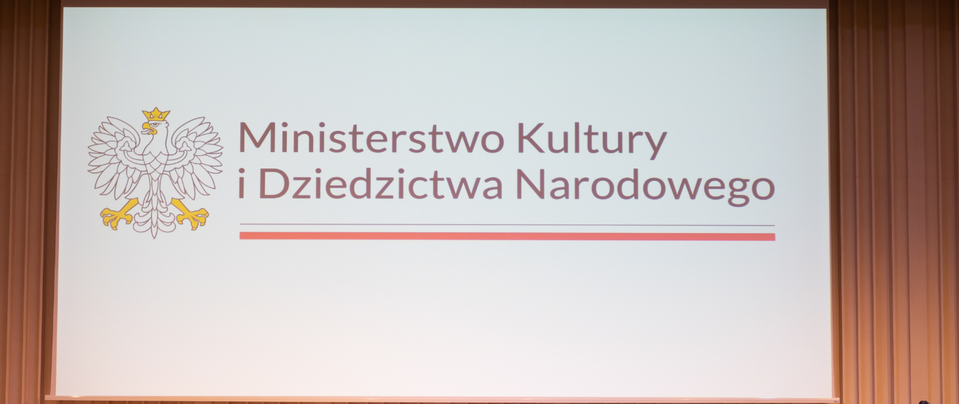 Na białym tle po lewej stronie biały orzeł w żółtej koronie. Po prawej stronie czarny napis Ministerstwo Kultury i Dziedzictwa Narodowego, a pod nim biało- czerwony pasek symbolizujący flagę Polski.