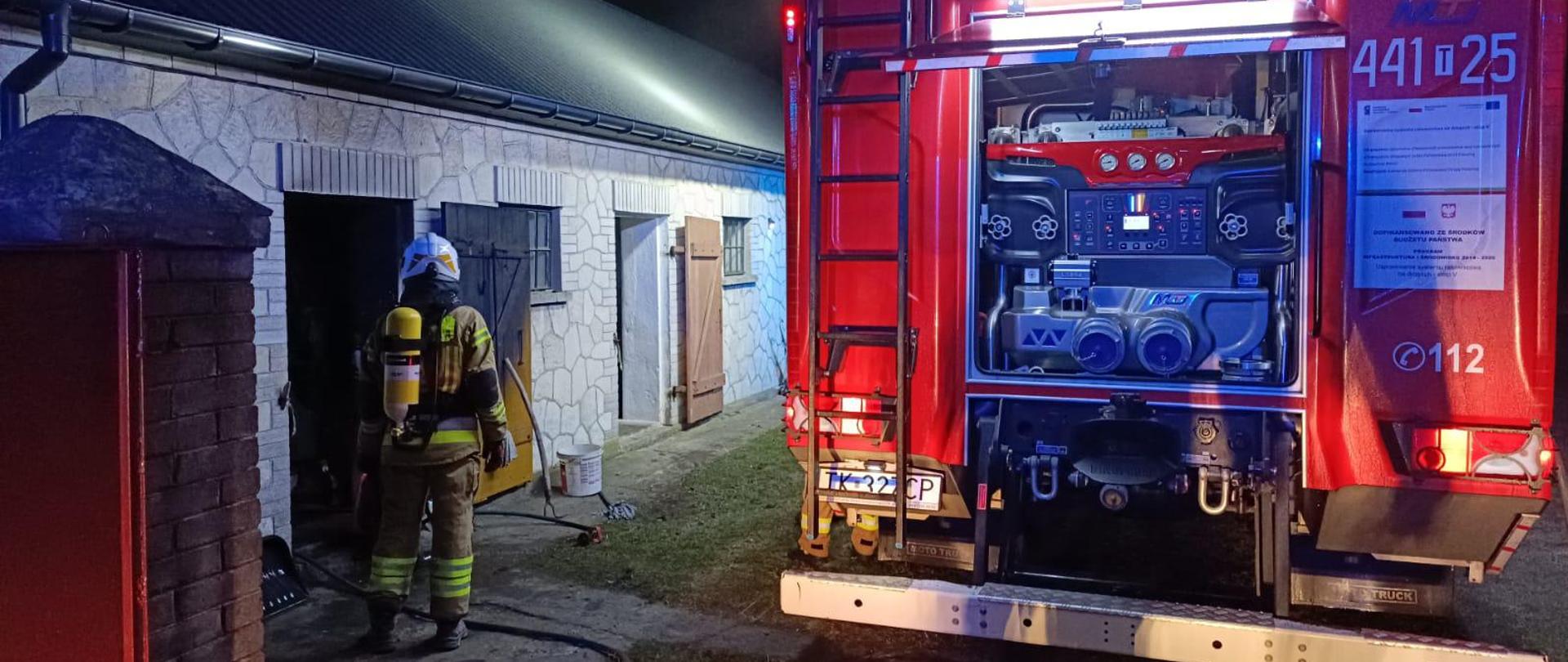 Nocna scena akcji strażackiej przy parterowym, murowanym budynku gospodarczym lub garażowym. Po lewej stronie zdjęcia widać strażaka ubranego w ubranie specjalne i aparatem oddechowym na plecach, stojącego przy otwartym, ciemnym wejściu do budynku. Na ziemi leży wąż strażacki prowadzący w stronę wejścia. Po prawej stronie stoi duży czerwony wóz strażacki z otwartą tylną skrytką, w której widać sprzęt i panel sterowania, intensywnie oświetlony białym oraz niebiesko-czerwonym światłem alarmowym. Ściana budynku jest jasna, pokryta dekoracyjnym wzorem przypominającym kamień, a kilka drzwi i okien jest otwartych. Cała scena sugeruje interwencję straży pożarnej podczas nocnego zdarzenia.