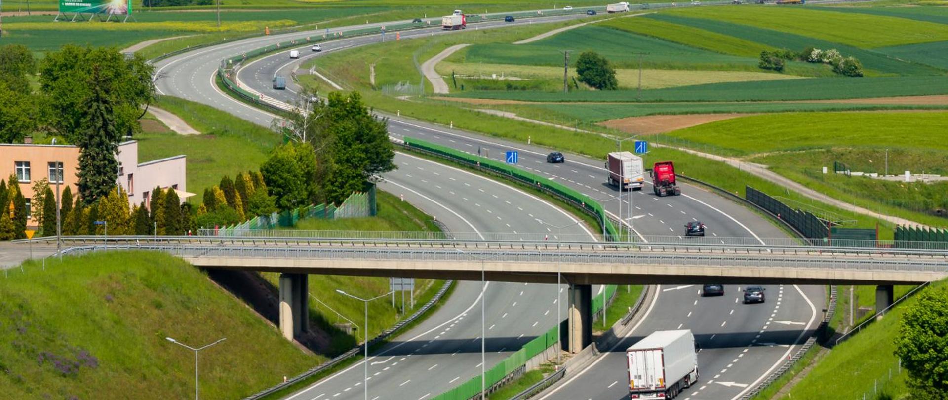 Autostrada A4 przebiegająca przez pagórkowaty krajobraz rolniczy. Na wielopasmowej jezdni widoczne są samochody osobowe i ciężarowe, a nad drogą przebiega wiadukt. Wokół rozciągają się zielone pola uprawne, trawniki oraz pojedyncze drzewa i zabudowania. Zdjęcie wykonane z podwyższonej perspektywy w słoneczny dzień.