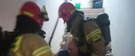 Zdjęcie przedstawia dwóch strażaków, którzy w czasie ćwiczeń ewakuują osobę poszkodowaną. Strażacy mają na sobie ubranie specjalne oraz aparaty powietrzne i maski. Zdjęcie wykonane jest na klatce schodowej.