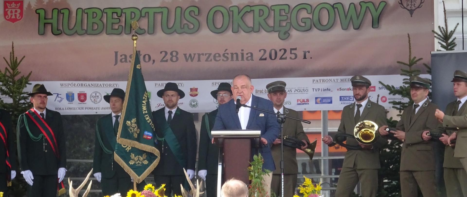 Mężczyzna w garniturze przemawia na scenie przy pulpicie, za nim stoi grupa mężczyzn ubranych w mundury służby leśnej z trąbkami w rękach. Po prawej stoi poczet ze sztandarem Polskiego Związku Łowieckiego.