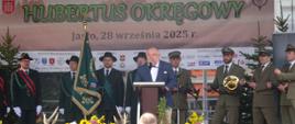Mężczyzna w garniturze przemawia na scenie przy pulpicie, za nim stoi grupa mężczyzn ubranych w mundury służby leśnej z trąbkami w rękach. Po prawej stoi poczet ze sztandarem Polskiego Związku Łowieckiego.