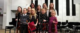 Na estradzie auli PSM przed fortepianem stoi w dwóch rzędach siedem dziewczynek i jeden chłopiec, wszyscy trzymają w rękach skrzypce i smyczki. Za nimi stoją dwie kobiety.