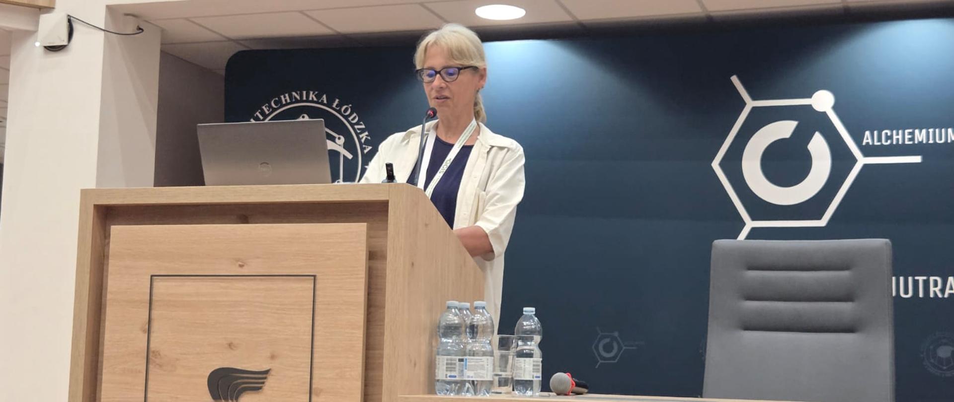 Aula. Za mównicą z mikrofonem stoi kobieta. Na mównicy logotyp Politechniki Łódzkiej. 