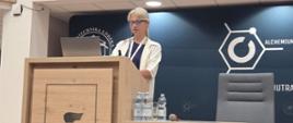Aula. Za mównicą z mikrofonem stoi kobieta. Na mównicy logotyp Politechniki Łódzkiej. 