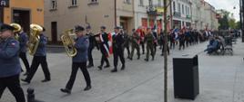 Narodowe Święto Trzeciego Maja. Rawicki rynek. Uczestnicy uroczystości maszerują w pochodzie patriotycznym. Tuż za orkiestrą - poczet sztandarowy z Komendy powiatowej PSP w Rawiczu. Za nim pozostałe poczty. Strażacy występują w mundurach galowych. W tle budynki.