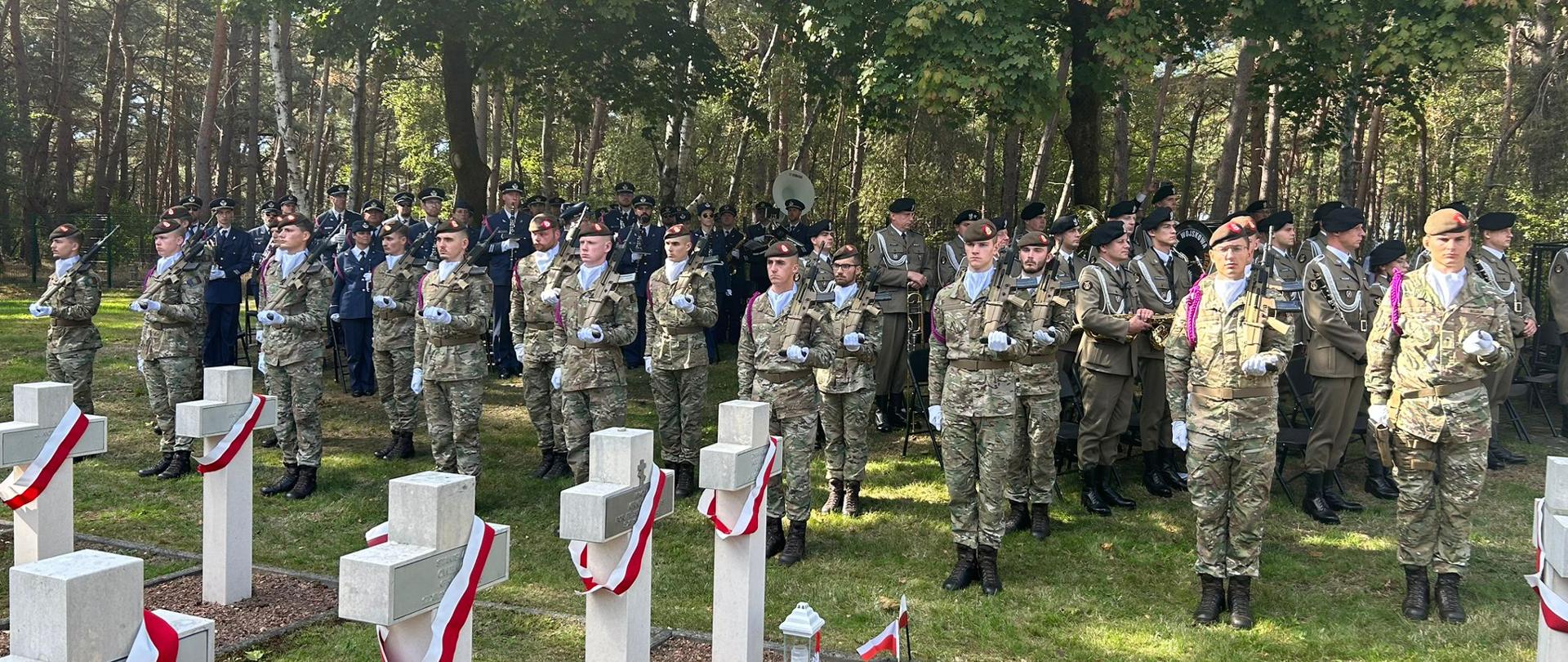 Guard of honour and military band standing close to the cementary. In the foreground crosses with a white and red sash.