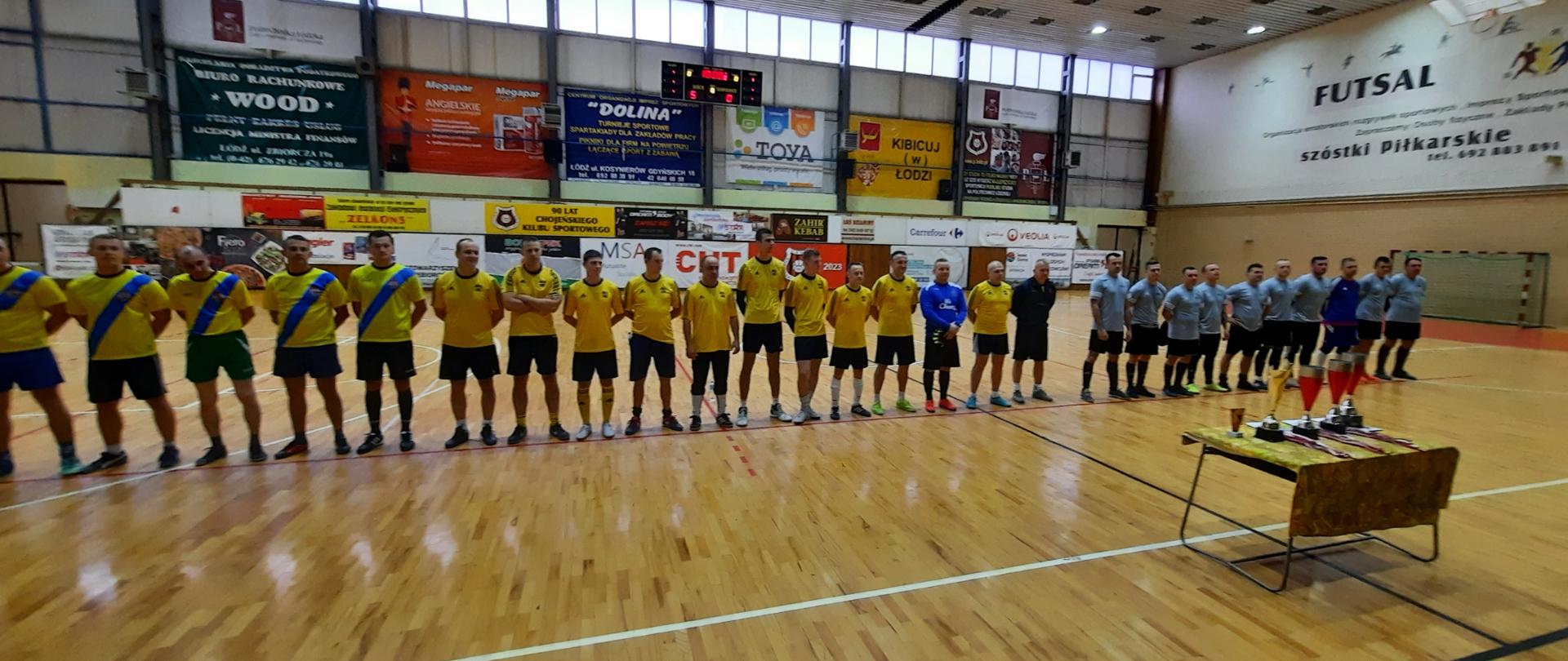 Strażacy w strojach sportowych ustawieni w rzędzie w oczekiwaniu na wręczenie nagród. Przed nimi stół z pucharami i medalami.