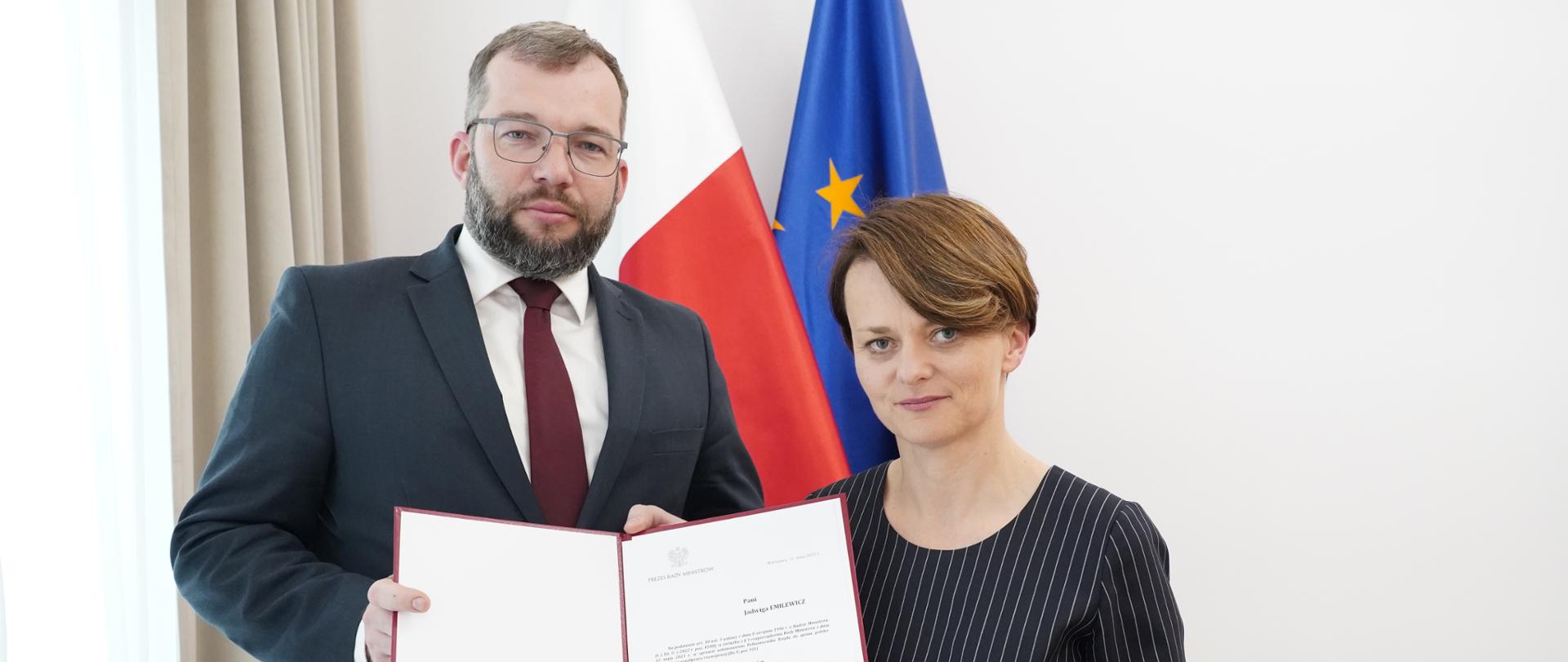 Minister Grzegorz Puda i wiceminister Jadwiga Emilewicz trzymają w rękach akt powołania - dokument w bordowej teczce. W tle flagi Polski i Unii Europejskiej.