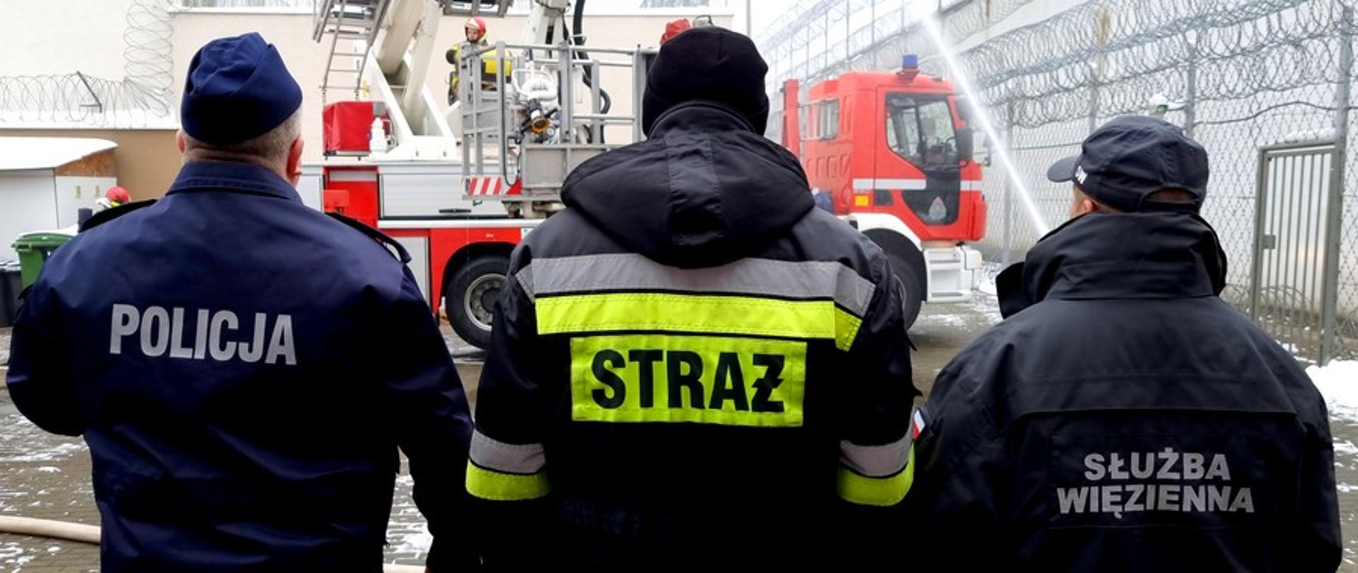 Zdjęcie przedstawia 3 funkcjonariuszy współpracujących służb. Widok z tyłu na napisy. W tle budynki i podnośnik strażacki.