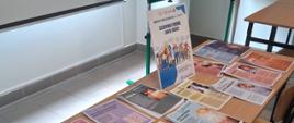 Zdjęcie przedstawia stół ustawiony w sali dydaktycznej, na którym rozłożone są materiały informacyjno edukacyjne dotyczące chorób zakaźnych i szczepień ochronnych. Na blatach leżą liczne ulotki i broszury, m.in. dot. odry, różyczki, RSV, krztuśca, tężca, inwazyjnej choroby pneumokokowej oraz zapobieganiu chorobom zakaźnym. Na środku stołu stoi plakat informacyjny poświęcony Europejskiemu Tygodniowi Szczepień.