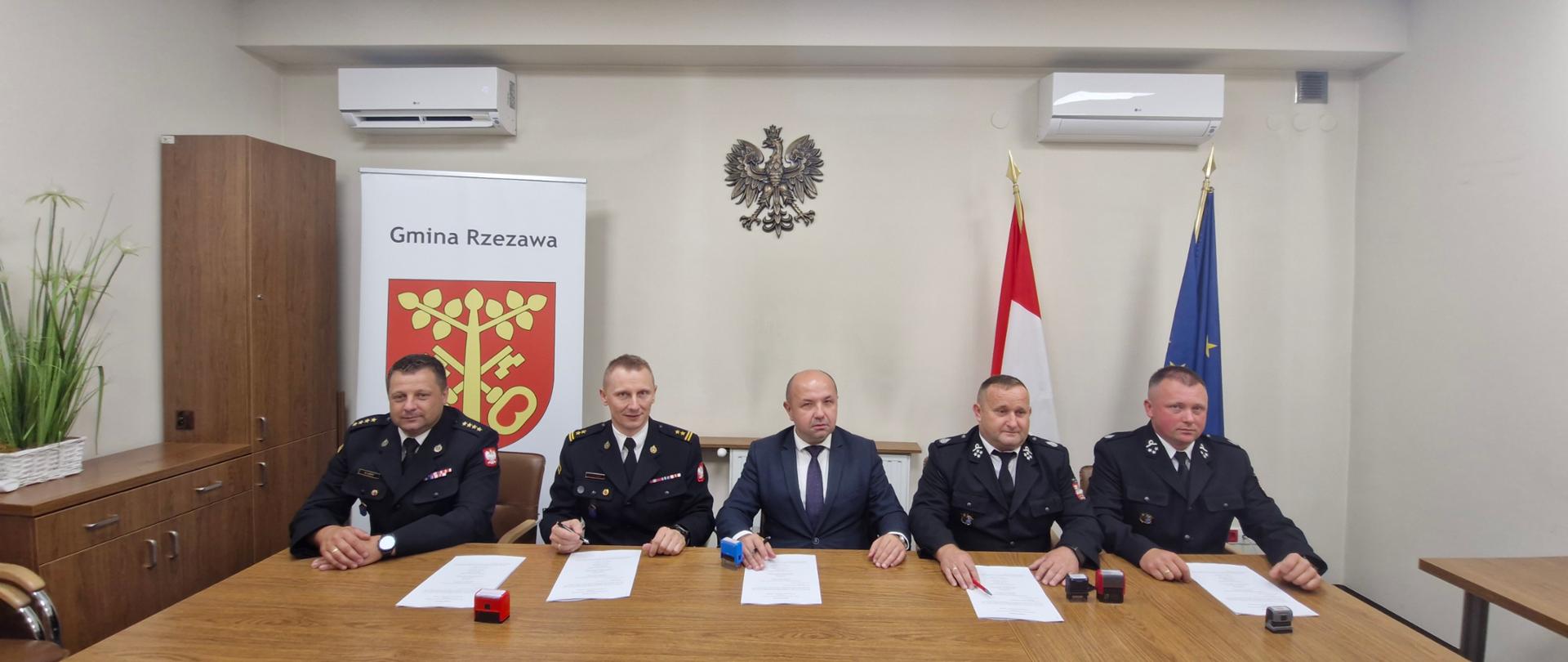 Pięć osób siedzi za drewnianym stołem. Czterech mężczyzn w mundurach strażackich i pomiędzy nimi jeden w eleganckim garniturze. Każdy z nich ma przed sobą dokumenty i pieczątki. W tle, po lewej stronie widać herb gminy Rzezawa, na środku, nad mężczyznami wisi godło Polski. Po prawej stronie znajduję się biało-czerwona flaga Polski oraz flaga Unii Europejskiej. 
