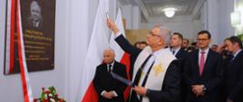 Widok na korytarz, na białej ścianie wisi ciemnoszara tablica z portretem Lecha Kaczyńskiego, przed nią stoi ksiądz trzymający w podniesionej ręce mały metalowy przedmiot, za nim grupa mężczyzn w garniturach.