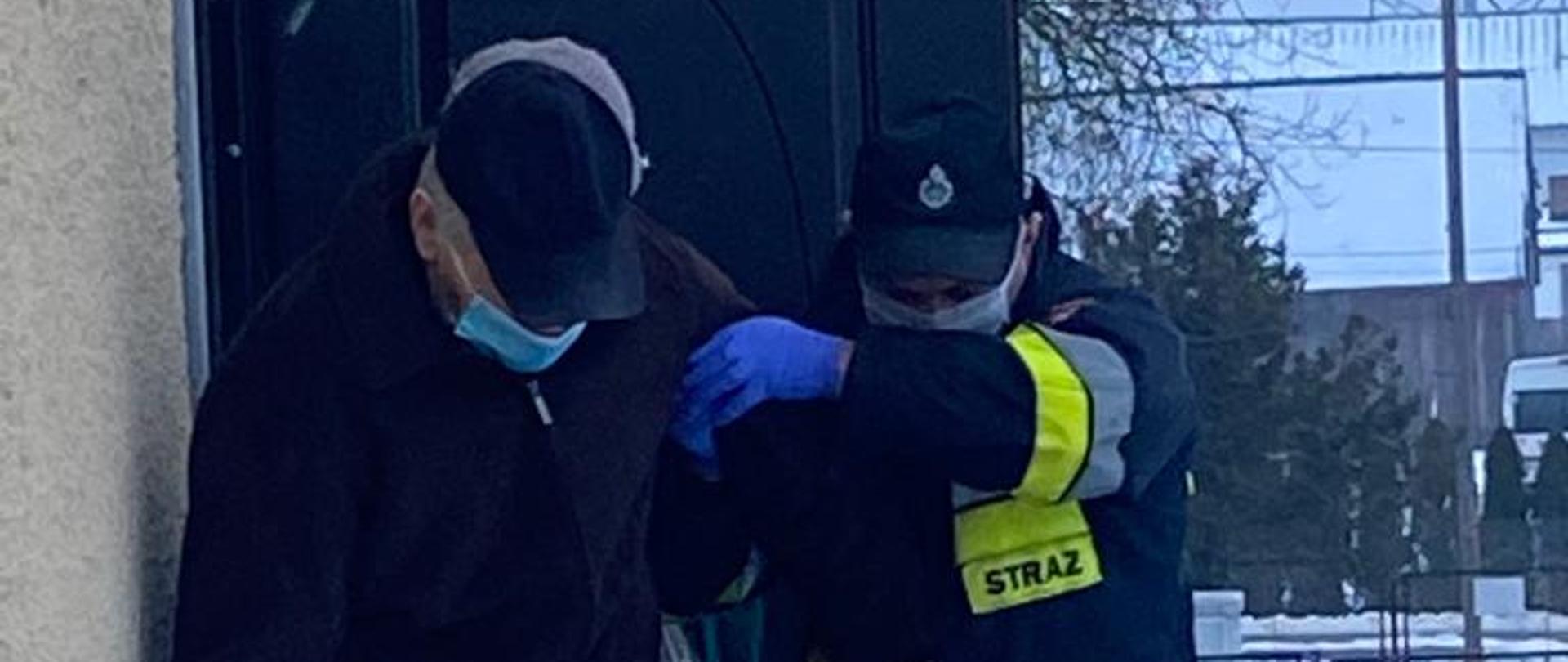 W centrum widać strażaka ubranego w ubranie specjalne, rękawiczki lateksowe i maseczkę. Strażak pomaga osobie starszej, poruszającej się o lasce zejść ze schodów.
