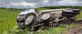 Przewrócona ciężarówka leżąca na boku w rowie. Pojazd leży na prawy boku, na pierwszym planie widoczne są koła lewej strony.