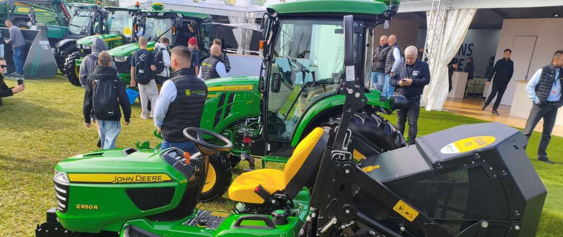"Zdjęcie przedstawia zielony traktorek ogrodowy (kosiarkę samojezdną) marki John Deere X950R, zaprezentowany na wystawie plenerowej. Maszyna stoi na trawie, zwrócona w lewą stronę, a w tle widać inne ciągniki rolnicze oraz zwiedzających ludzi.
Oto szczegółowy opis kluczowych elementów pojazdu:
• Nadwozie i przód: Traktorek ma charakterystyczną dla marki John Deere żywozieloną maskę z żółtym napisem „JOHN DEERE” oraz mniejszym, czerwono-białym oznaczeniem modelu „X950R” na boku. Przód maski wieńczy srebrna osłona chłodnicy z reflektorami.
• Koła: Pojazd wyposażony jest w cztery szerokie, czarne opony z głębokim bieżnikiem trawnikowym, zamontowane na jaskrawożółtych felgach. Przednie koła są wyraźnie mniejsze od tylnych.
• Agregat tnący: Podwozie traktorka, pomiędzy przednią a tylną osią, zajmuje zielona obudowa noży tnących (kosisko) z dodatkowymi małymi, czarnymi kółkami podporowymi zapobiegającymi uszkodzeniom darni.
• Miejsce operatora: Na środku znajduje się odsłonięte, ergonomiczne, wysokie siedzisko w kolorze żółtym. Przed nim umieszczono czarną kierownicę oraz deskę rozdzielczą. Po prawej stronie fotela wznosi się wysoki, czarny pałąk ochronny (ROPS).
• Tył pojazdu: Do tylnej części traktorka przymocowany jest bardzo duży, czarny, zabudowany kosz na trawę z metalowym stelażem i hydraulicznym systemem unoszenia, służący do zbierania i wysokiego opróżniania pokosu."