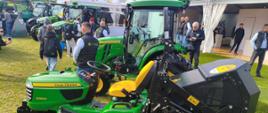 "Zdjęcie przedstawia zielony traktorek ogrodowy (kosiarkę samojezdną) marki John Deere X950R, zaprezentowany na wystawie plenerowej. Maszyna stoi na trawie, zwrócona w lewą stronę, a w tle widać inne ciągniki rolnicze oraz zwiedzających ludzi.
Oto szczegółowy opis kluczowych elementów pojazdu:
• Nadwozie i przód: Traktorek ma charakterystyczną dla marki John Deere żywozieloną maskę z żółtym napisem „JOHN DEERE” oraz mniejszym, czerwono-białym oznaczeniem modelu „X950R” na boku. Przód maski wieńczy srebrna osłona chłodnicy z reflektorami.
• Koła: Pojazd wyposażony jest w cztery szerokie, czarne opony z głębokim bieżnikiem trawnikowym, zamontowane na jaskrawożółtych felgach. Przednie koła są wyraźnie mniejsze od tylnych.
• Agregat tnący: Podwozie traktorka, pomiędzy przednią a tylną osią, zajmuje zielona obudowa noży tnących (kosisko) z dodatkowymi małymi, czarnymi kółkami podporowymi zapobiegającymi uszkodzeniom darni.
• Miejsce operatora: Na środku znajduje się odsłonięte, ergonomiczne, wysokie siedzisko w kolorze żółtym. Przed nim umieszczono czarną kierownicę oraz deskę rozdzielczą. Po prawej stronie fotela wznosi się wysoki, czarny pałąk ochronny (ROPS).
• Tył pojazdu: Do tylnej części traktorka przymocowany jest bardzo duży, czarny, zabudowany kosz na trawę z metalowym stelażem i hydraulicznym systemem unoszenia, służący do zbierania i wysokiego opróżniania pokosu."