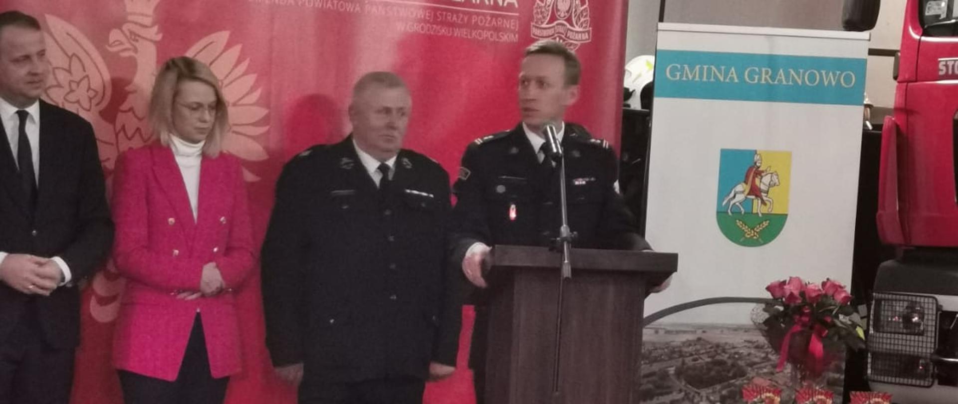 strażak stojący przy mównicy, za nim stoją w szeregu trzy osoby, strażack, kobieta w czarnych spodniach i różowej marynarce, mężczyzna w garniturze, za nimi czerwony banerz orłem