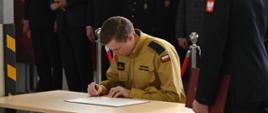 Strażak składa podpis pod dokumentami potwierdzającymi jego przyjęcie do służby, w tle zaproszeni goście