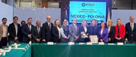 En la foto se ve a un grupo de unas quince personas de pie en semicírculo detrás de una larga mesa cubierta con un mantel verde. Todos van elegantemente vestidos, con trajes, chaquetas y atuendos formales, y miran hacia la cámara, posando para una foto conjunta.
En el centro de la mesa, delante del grupo, hay dos obsequios que simbolizan a Polonia y México: un libro blanco y rojo, un álbum con la inscripción «Poland», y una vaza decorativa con motivos folclóricos mexicanos. En medio del grupo se encuentra la embajadora de Polonia en México, Agnieszka Frydrychowicz-Tekieli, sosteniendo uno de los obsequios.
Detrás de los participantes se ve una gran pantalla con la inscripción:
«Cámara de Diputados – LXVI Legislatura. Instalación Grupo de Amistad México–Polonia. Palacio Legislativo de San Lázaro, 21 de octubre de 2025».
A los lados de la pantalla hay banderas de México y Polonia.
La foto fue tomada en la sala de conferencias de la Cámara de Diputados de México. El interior es moderno, con paredes claras y techo de madera. La imagen documenta el momento oficial de la instalación del Grupo de Amistad México-Polonia, un evento de carácter solemne y diplomático, lleno de cordialidad y cooperación entre los participantes.