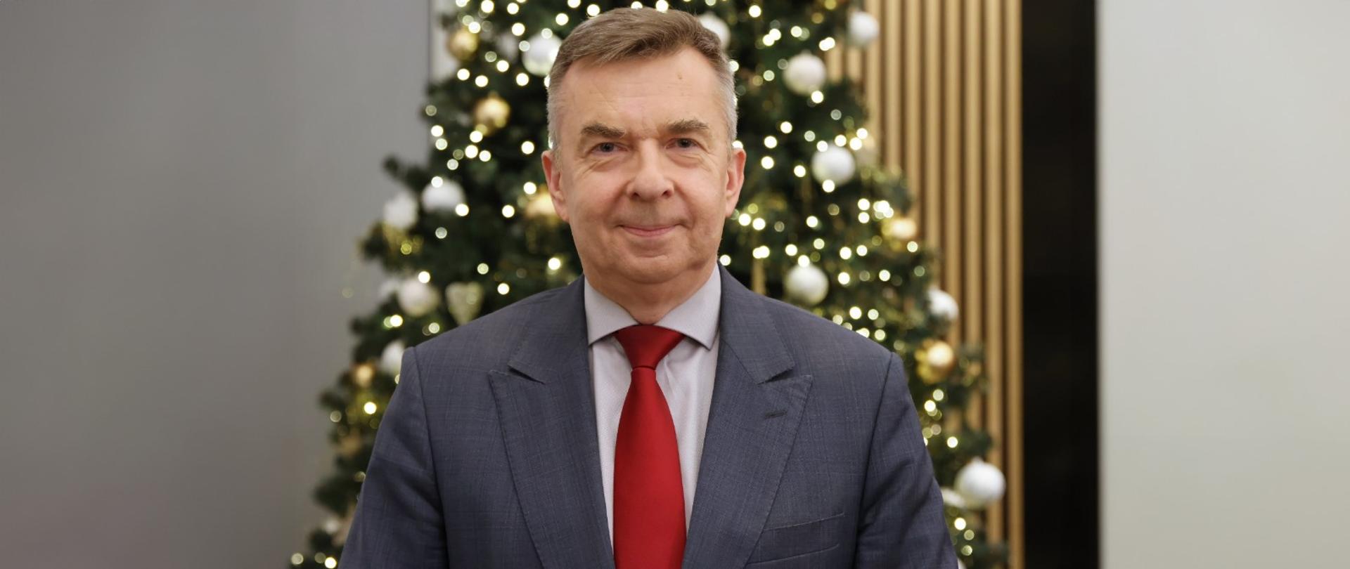 Minister Wieczorek stoi na tle udekorowanej bombkami i łańcuchami choinki.