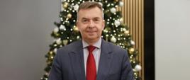 Minister Wieczorek stoi na tle udekorowanej bombkami i łańcuchami choinki.