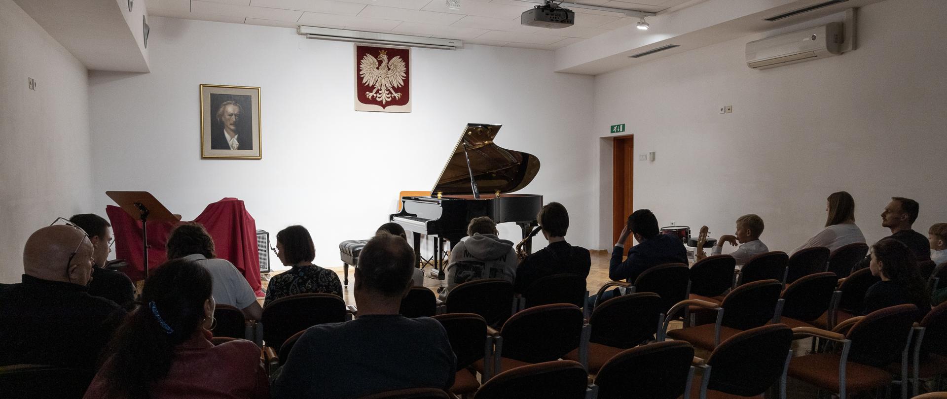 scena auli w państwowej szkole muzycznej w głubczycach, na scenie otwarty fortepian i zestaw perkusyjny zasłonięty czerwoną płachtą, na pierwszym planie osoby siedzące na publiczności