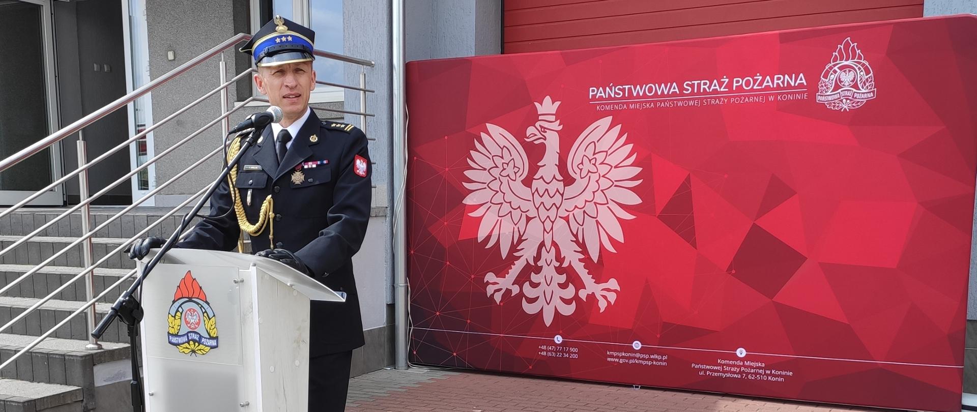strażak stoi przed mównicą, za nim czerwona ścianka oraz budynek