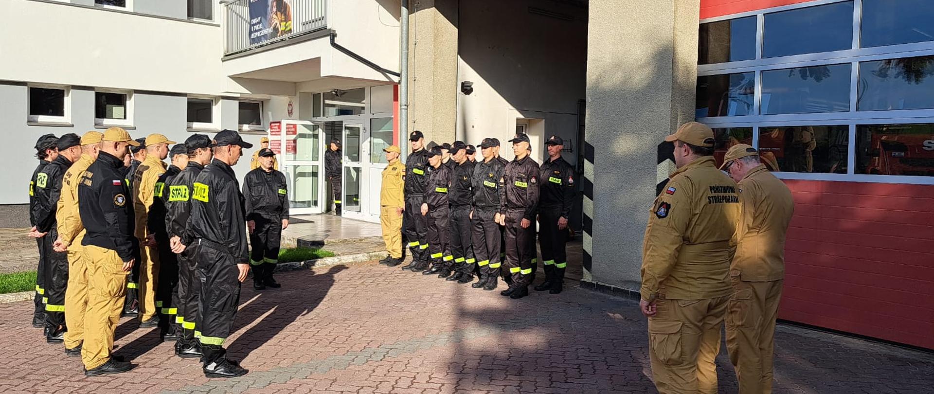 Ujęcie z boku na ustawionych w szeregu strażaków PSP w czarnych mundurach stojących pod budynkiem remizy; naprzeciwko nich dowódcy PSP w jasnych strojach, w tle wejście do budynku i baner promujący bezpieczeństwo.