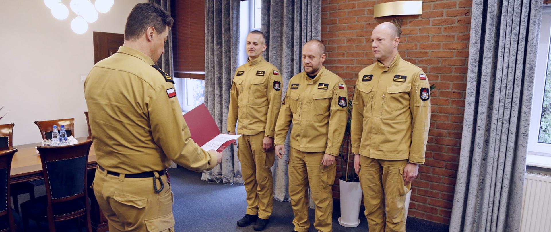 Gabinet Komendanta CS PSP, trzech funkcjonariuszy CS PSP stojących w szeregu, przed nimi stojący Komendant Centralnej Szkoły PSP trzymający w dłoniach czerwoną teczkę z pismem, które odczytuje