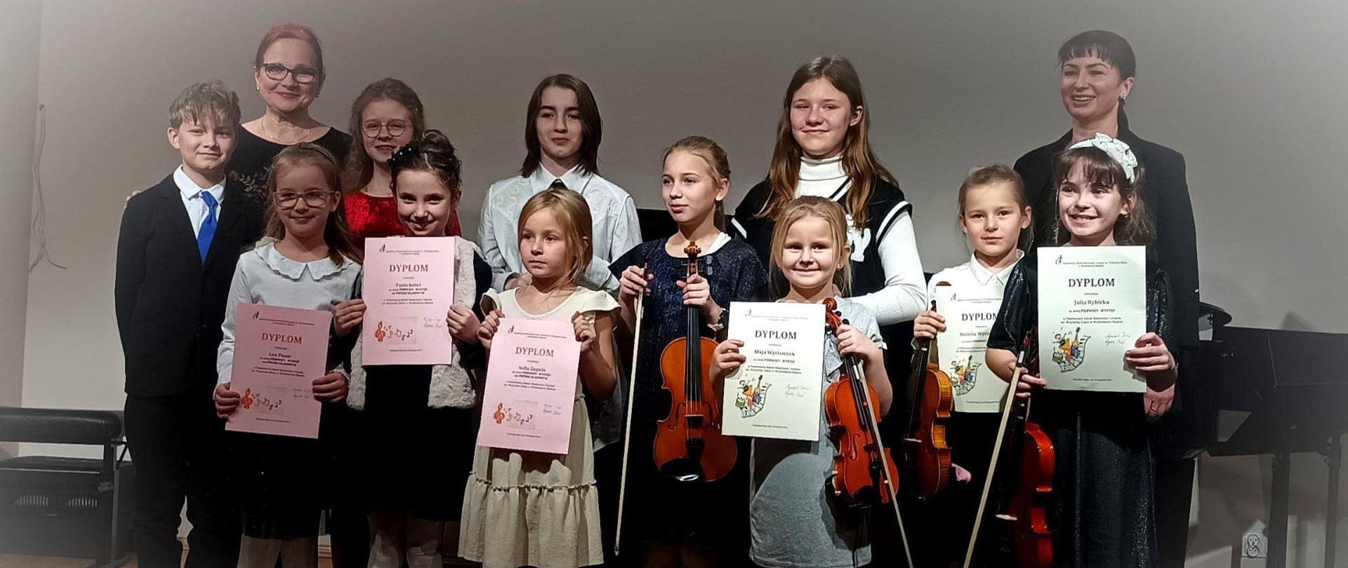 Grupa uczniów wraz z dwoma nauczycielkami stoi na scenie, trzymają w rękach skrzypce i dyplomy