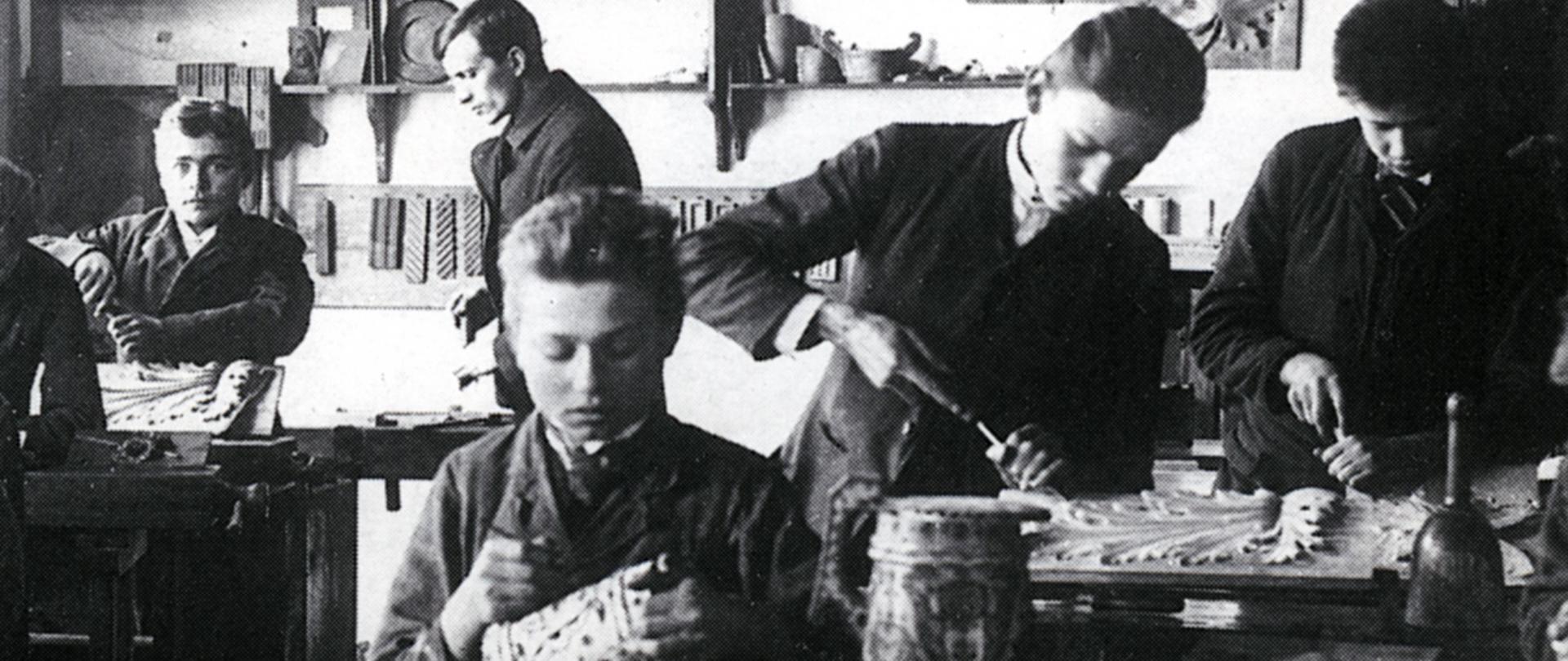 Na czarno-białym zdjęciu widzimy wnętrze pracowni rzeźby. Fotografia została zrobiona podczas zajęć praktycznych z rzeźby. Na pierwszym planie widzimy ucznia siedzącego przy warsztacie stolarskim i pracującego nad drewnianym pucharem. Uczeń siedząc przy warsztacie trzyma w rękach drewniany puchar, na którym coś rysuje. Przed uczniem na warsztacie stoi drugi taki sam puchar. Na drugim planie widzimy jeszcze czterech innych uczniów rzeźbiących przy warsztatach stolarskich. Ci uczniowie rzeźbią motywy roślinne. Z tyłu w tle na białej ścianie widzimy ekspozycję prac uczniowski: drewniane rzeźby, talerze, studium ręki, motywy roślinne i ptaki. Dodatkowo na ścianie widzimy oprawiony portret Cesarza Franciszka Józefa.