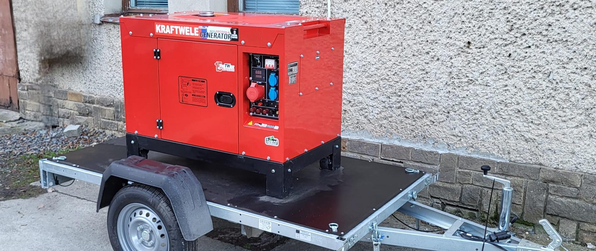 Na zdjęciu widoczny jest czerwony generator prądu marki KRAFTWELE GENERATOR, zamontowany na małej, jednoosiowej przyczepie transportowej. Generator ma widoczny panel sterowania z wskaźnikami, różnymi gniazdami elektrycznymi (niebieskie i czerwone gniazda przemysłowe, standardowe gniazda domowe) oraz czerwony przycisk awaryjnego zatrzymania. Jest on osadzony na czarnych metalowych nogach, przymocowanych do czarnej, drewnianej platformy przyczepy. Przyczepa ma srebrną ramę, jedno koło z błotnikiem, zaczep kulowy i koło podporowe. Tło stanowi jasna, teksturowana ściana budynku z oknem z żaluzjami oraz betonowe podłoże.