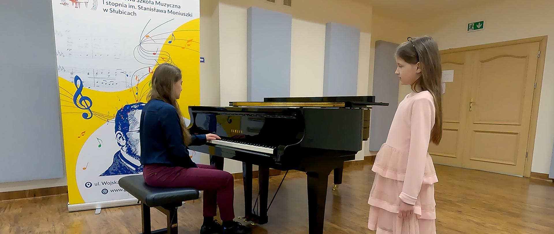 W auli szkolnej nauczycielka gra na fortepianie, obok niej stoi uczennica. 