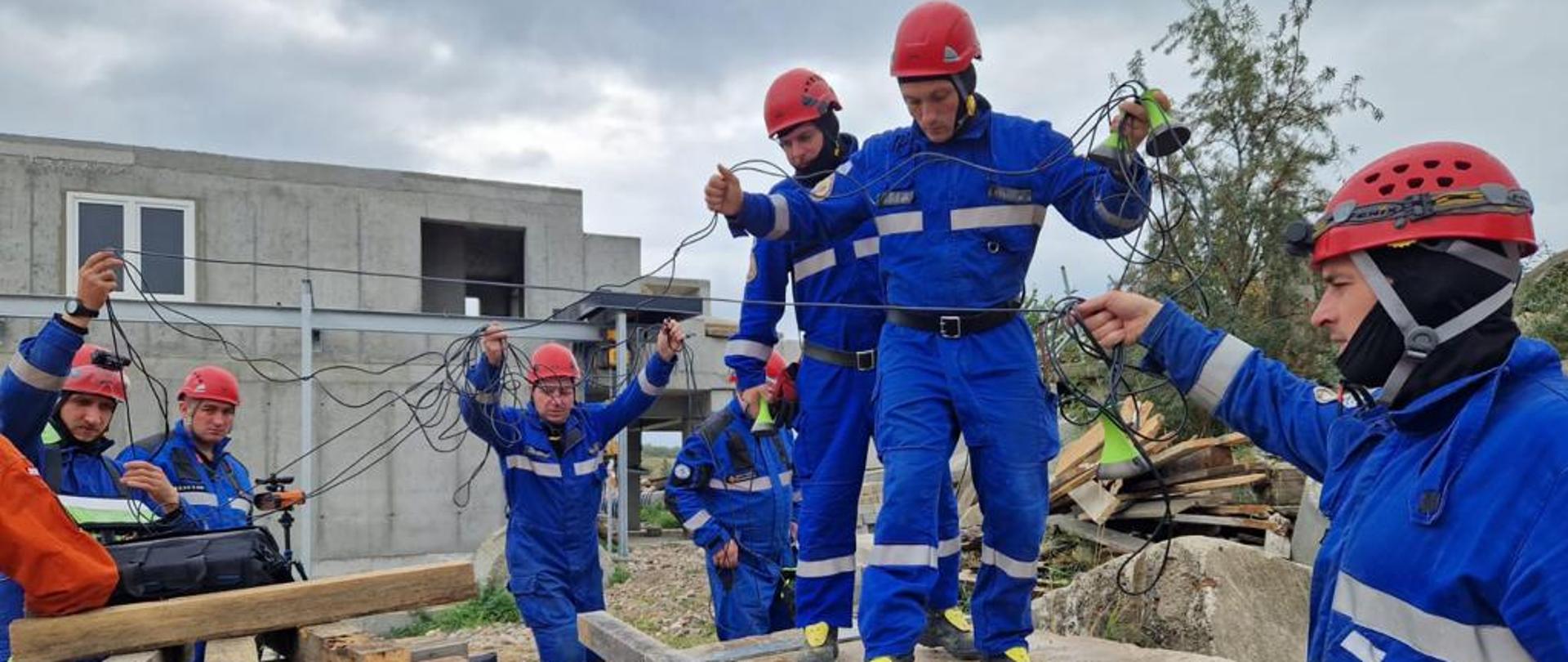 Grupa mężczyzn w niebieskich kombinezonach i kaskach stoi wokół i na płytach cementowych i rozciąga kable podłączone do sprzętu ratowniczego