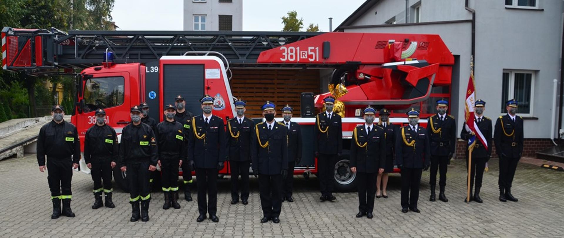 Strażacy ustawieni w luźnym szyku, stoją przed drabiną samochodową, po prawej stronie widoczny jest poczet sztandarowy składający się z trzech strażaków, w centrum fotografii widoczny jest strażak odchodzący na emeryturę - Marin Kosior. Część strażaków występuje w umundurowaniu galowym, a część w umundurowaniu koszarowym. Po prawej stronie w tle oraz w oddali widoczny jest budynek komendy.