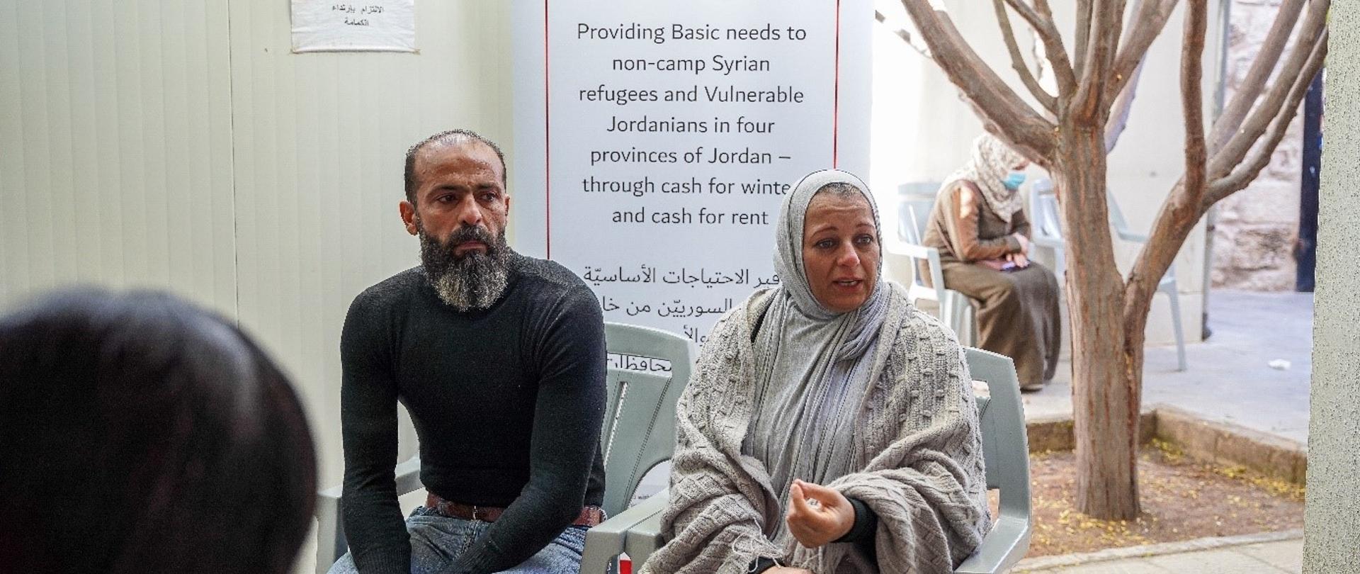 Mężczyzna z zarostem w czarnej bluzce i kobieta z głową i ramionami okrytymi chustami siedzą na krzesłach. W tle znajduje się plakat z logo Polskiej pomocy i Caritas