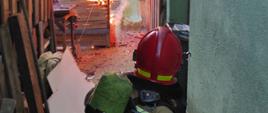 Na zdjęciu widać strażaka przy elewacji budynku mieszkalnego oraz płomienie wydostające się z budynku. 