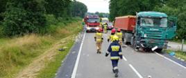 Na zdjęciu widać samochód ciężarowy z naczępą po zderzeniu z samochodem dostawczym- śmieciarką, na drodze rozsypane śmieci, za samochodem cięzarowym samochód ratowniczo-gaśniczy OSP Szczucin i Zespół Ratownictwa Medycznego, droga krajowa zablokowana, na drodze strażacy udający się na rozpoznanie.