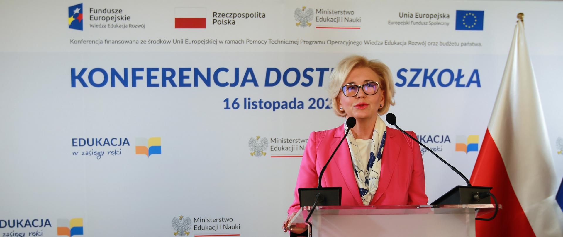 Wiceminister Machałek stoi przy mównicy i mówi do mikrofonu, za nią na ścianie napis Konferencja dostępna szkoła - 16 listopada 2022 r.