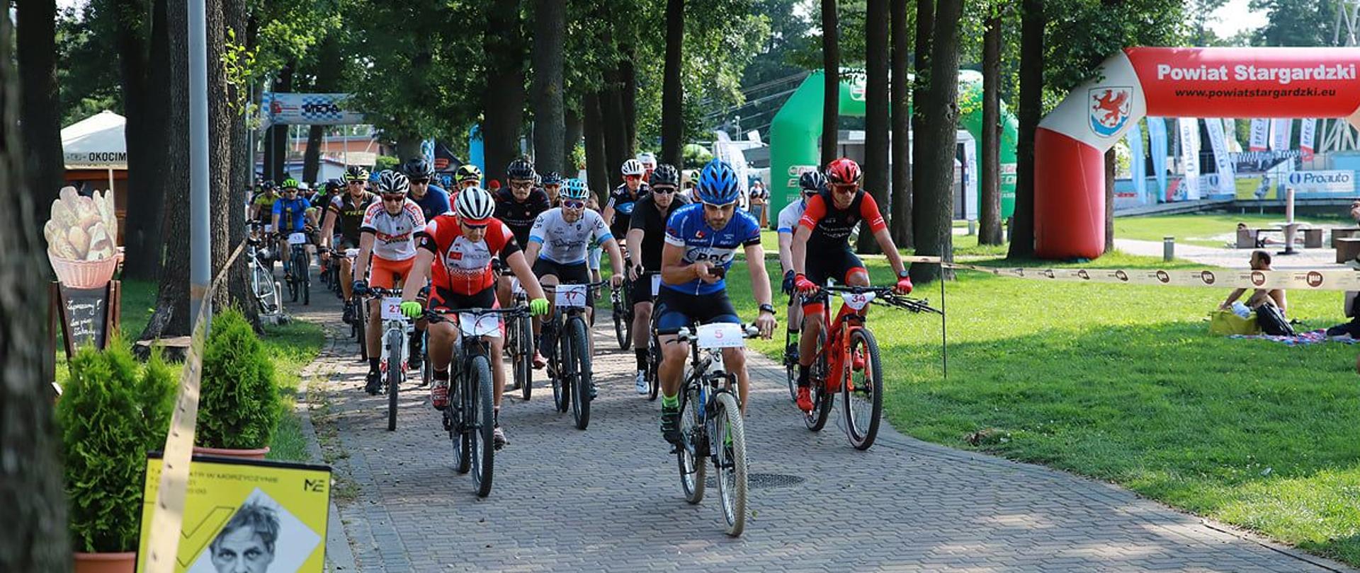 Na zdjęciu widoczni są uczestnicy zawodów MTB poruszający się trasą wyścigu po kostce brukowej. W tle widoczny park wraz z reklamami Powiatu Stargardzkiego, widownia siedząca na trawie oraz elementy charakterystyczne dla parku - drzewa, ławki, chodniki. 